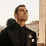 Valentino Garavani poses elegantly with silver brooch and fedora against red velvet backdrop