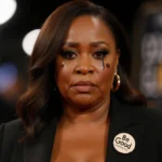 Wanda Sykes wearing Be Good pin with emotional expression and blurred red carpet behind