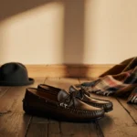 Leather loafers rest on worn wooden floor with winter accessories scattered nearby and warm beige wall behind