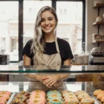 Young woman smiles behind cookie shop counter with colorful sprinkles and fresh treats in glass case (91 chars)