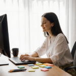Woman sitting at minimalist desk working on laptop with natural light pouring in and coffee beside her