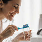Young woman rinsing toothbrush with faucet off and soap dispenser droplets in natural light
