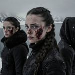 Three women stand on snowy peak with one determined gaze and another bloodied while misty frozen lake stretches behind them