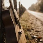 Worn wooden guitar leans against weathered fencepost with soft sunlight casting long shadows and distant road stretching behi