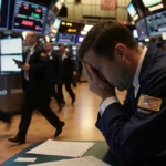 Worried stock market analyst hunched over computer screen with falling stock data and papers scattered on desk