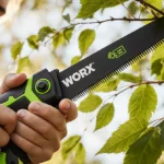 Person pruning tree branch with cordless pruning saw showing focused hands and greenery